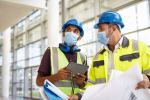 Two people in hardhats and masks looking at blueprints in a building